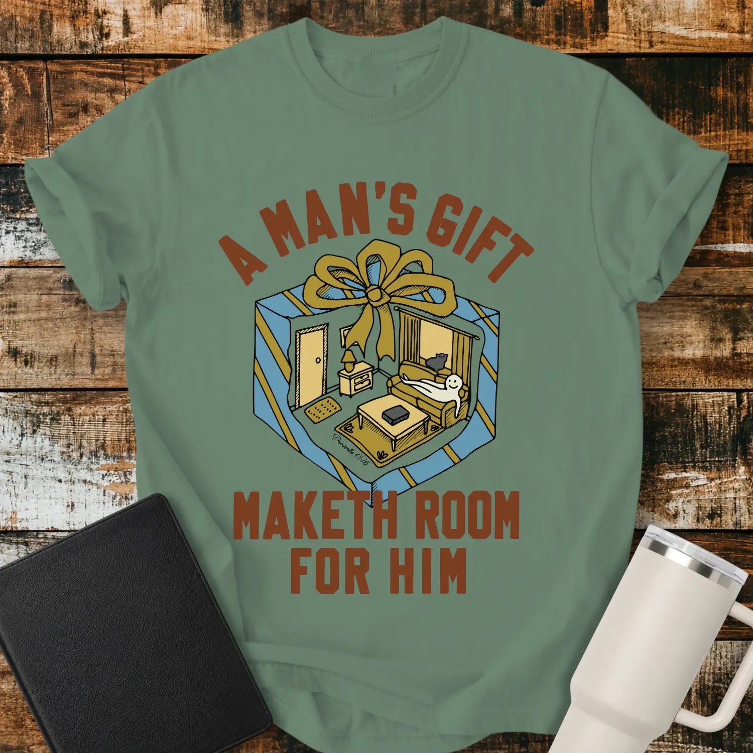 light green graphic T-shirt mockup featuring the phrase “A Man’s Gift Maketh Room for Him” in vintage-style lettering with an illustration of a gift box opening into a furnished room scene, styled with a black notebook and white tumbler on a rustic wood background.