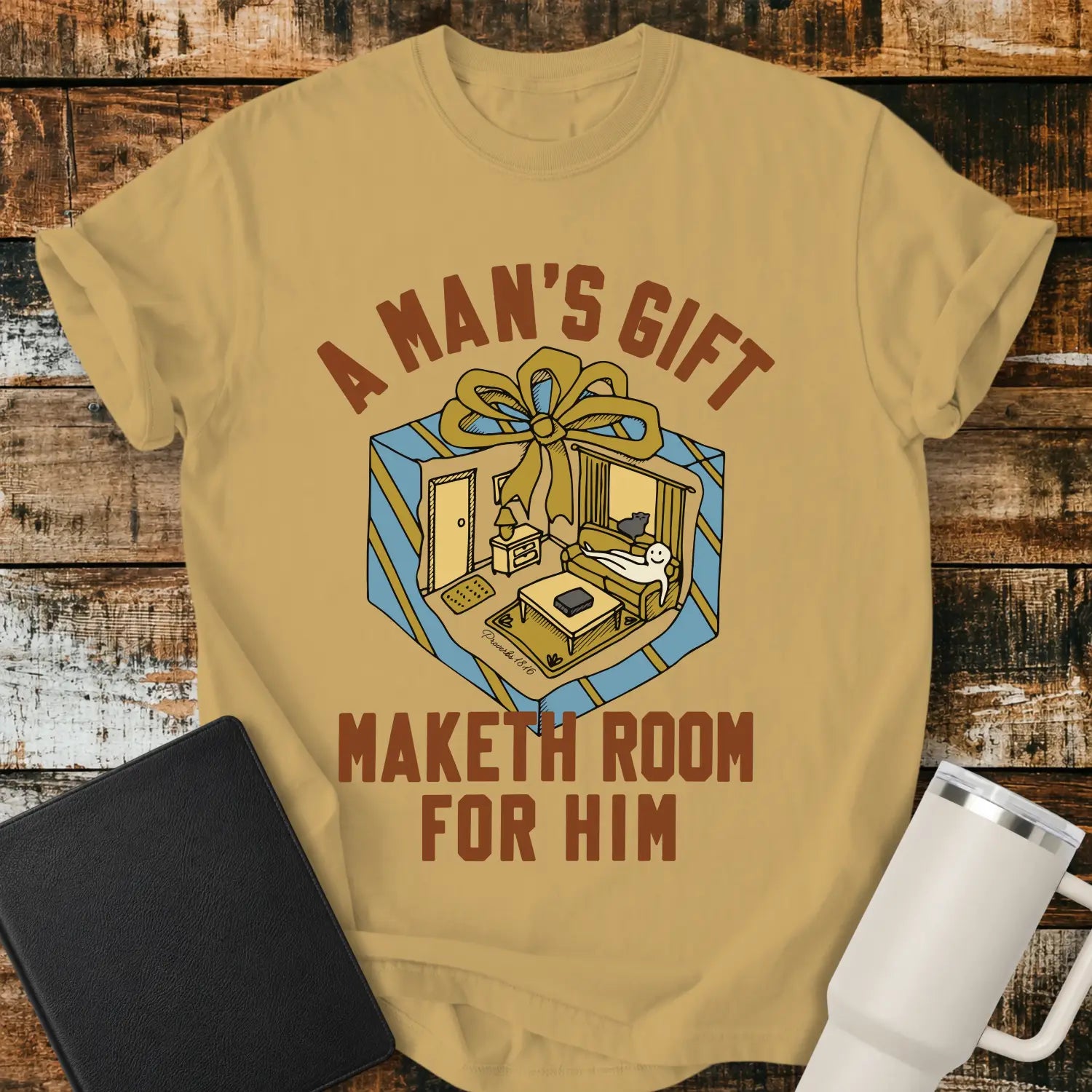 Mustard yellow graphic T-shirt mockup featuring the phrase “A Man’s Gift Maketh Room for Him” in vintage-style lettering with an illustration of a gift box opening into a furnished room scene, styled with a black notebook and white tumbler on a rustic wood background.