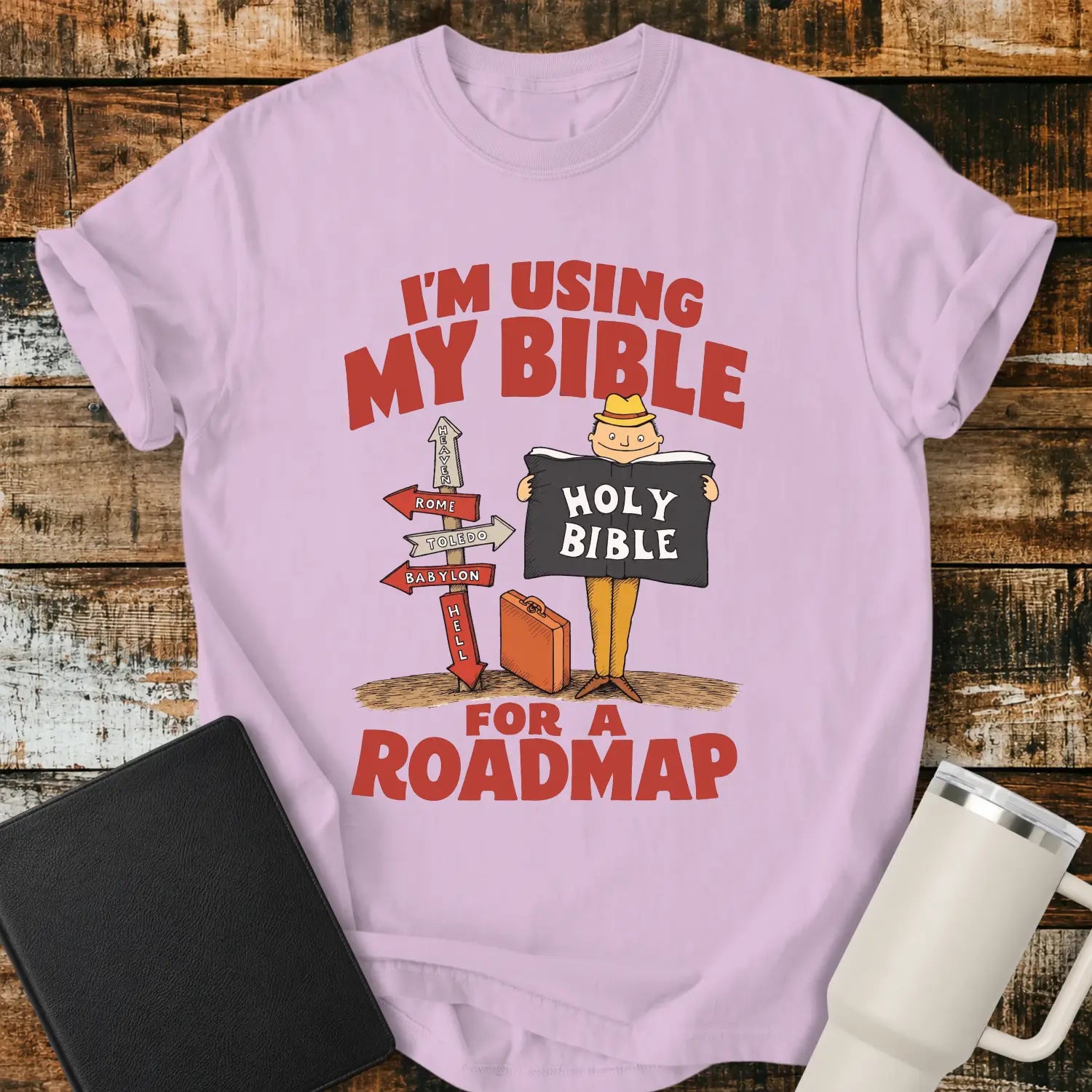 orchid purple graphic T-shirt mockup featuring the phrase “I’m Using My Bible for a Roadmap” in bold red lettering with a cartoon traveler holding a Holy Bible beside directional signs, styled with a black Bible and white tumbler on a rustic wood background.