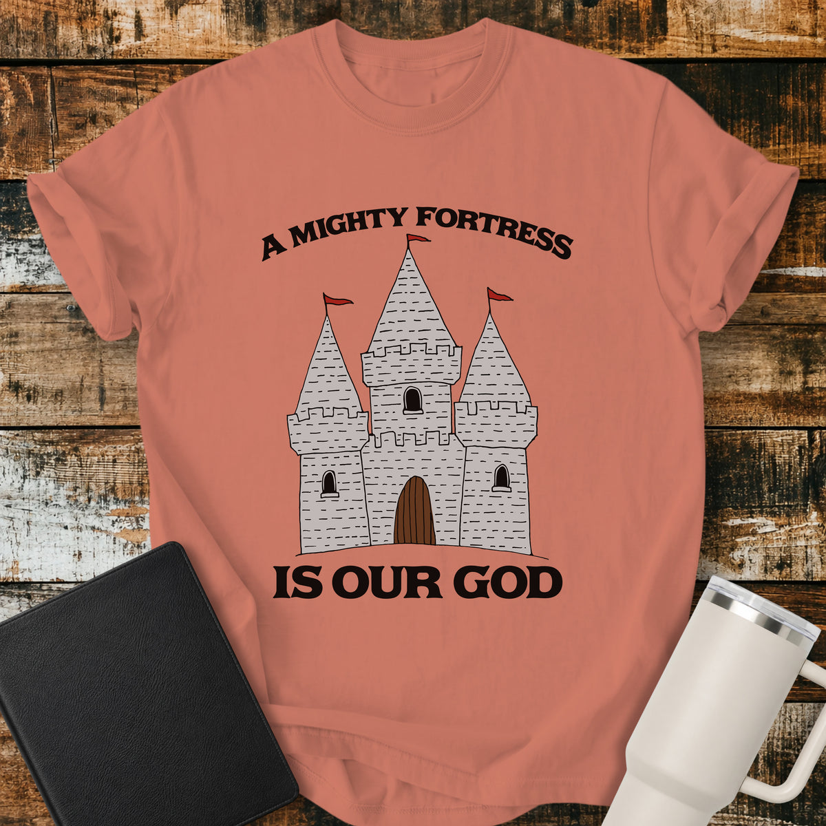 terracotta Christian t-shirt featuring a hand-drawn castle illustration with bold vintage lettering reading “A Mighty Fortress Is Our God.”