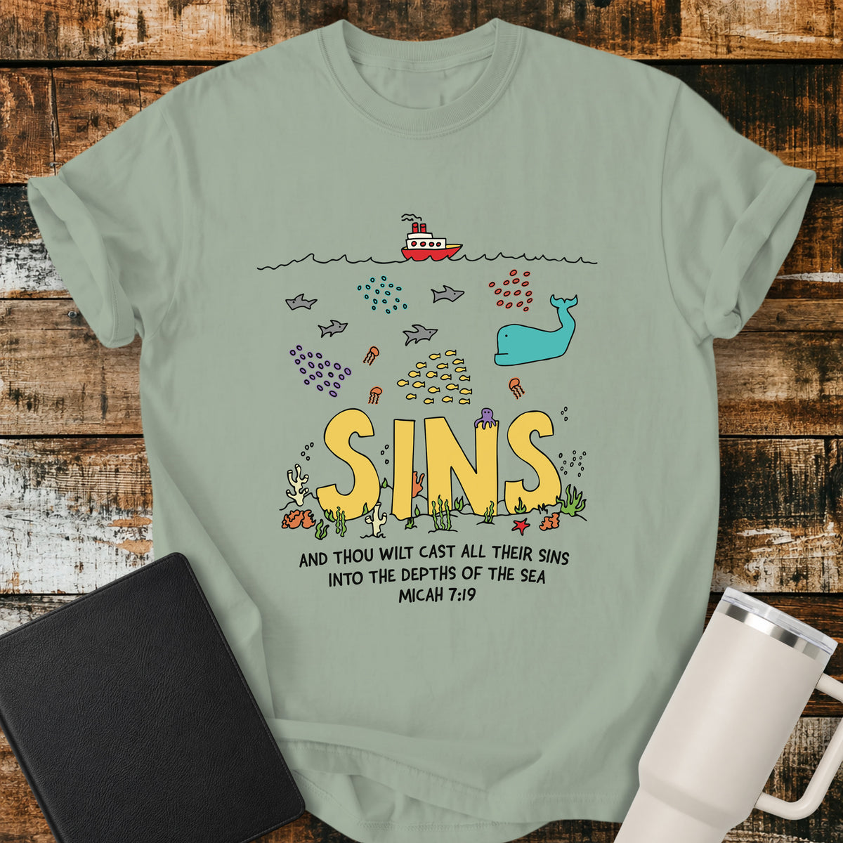bay green Christian t-shirt featuring a colorful undersea illustration with the word “SINS” resting on the ocean floor, surrounded by fish, a whale, coral, and sea life, with Micah 7:19 below: “And thou wilt cast all their sins into the depths of the sea.”