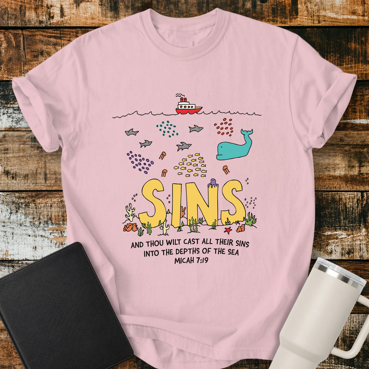 blossom pink Christian t-shirt featuring a colorful undersea illustration with the word “SINS” resting on the ocean floor, surrounded by fish, a whale, coral, and sea life, with Micah 7:19 below: “And thou wilt cast all their sins into the depths of the sea.”
