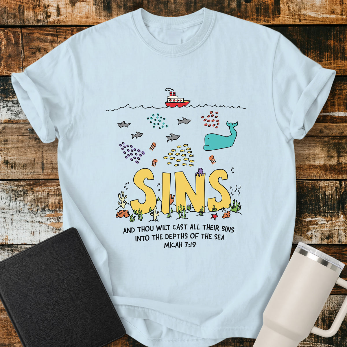 Chambray blue Christian t-shirt featuring a colorful undersea illustration with the word “SINS” resting on the ocean floor, surrounded by fish, a whale, coral, and sea life, with Micah 7:19 below: “And thou wilt cast all their sins into the depths of the sea.”