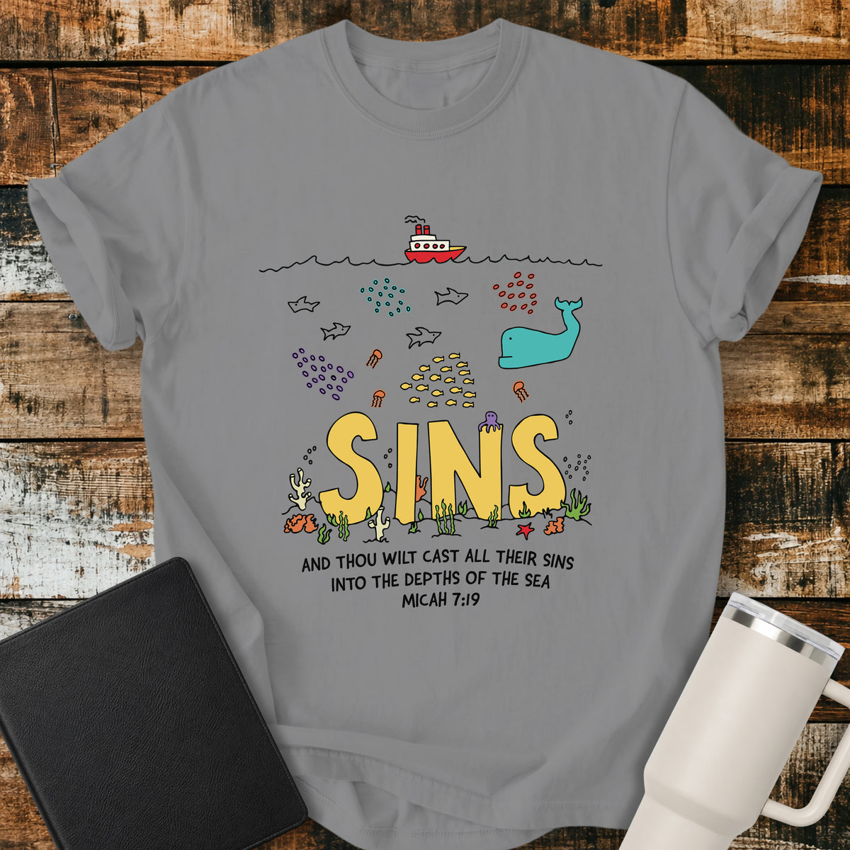 grey Christian t-shirt featuring a colorful undersea illustration with the word “SINS” resting on the ocean floor, surrounded by fish, a whale, coral, and sea life, with Micah 7:19 below: “And thou wilt cast all their sins into the depths of the sea.”