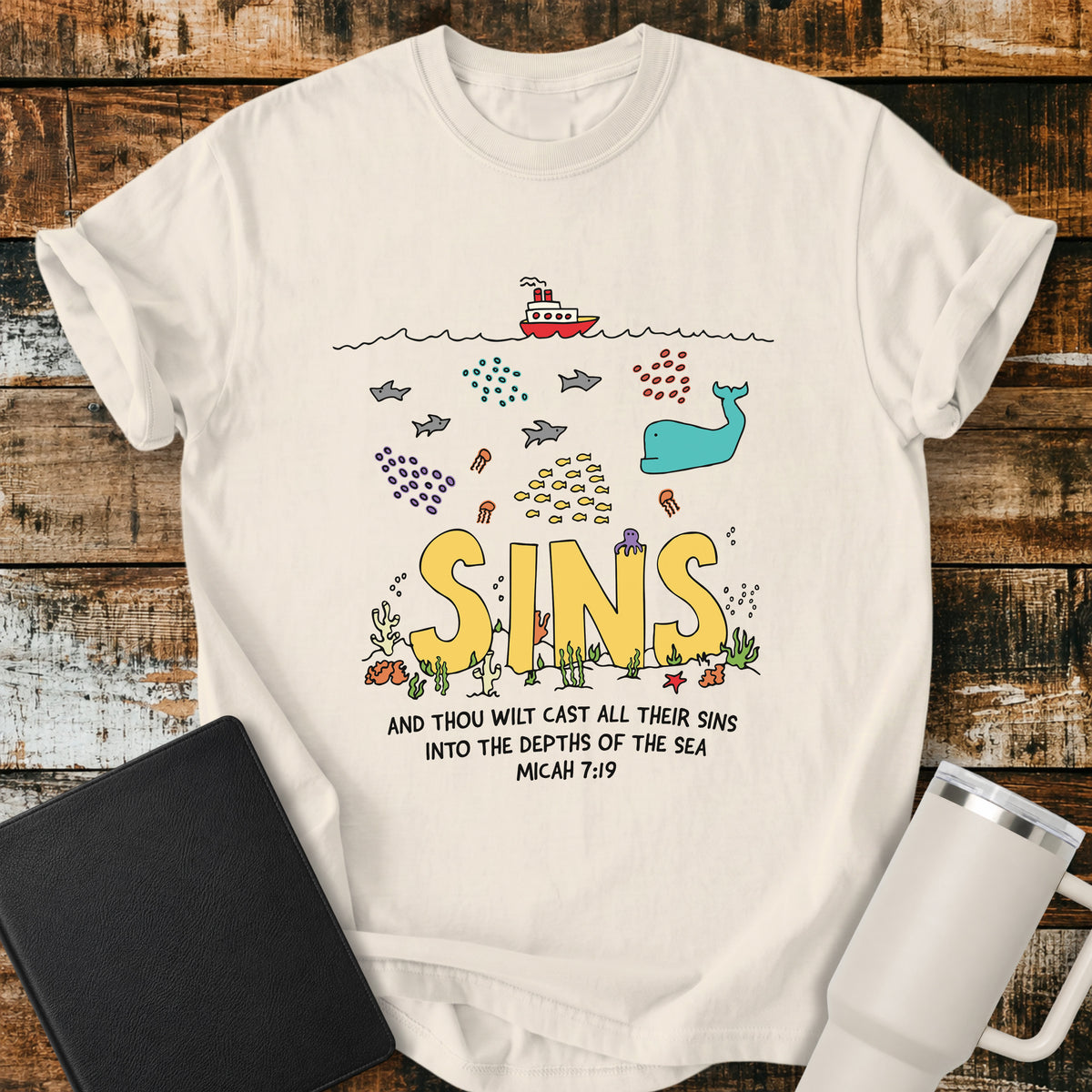 ivory Christian t-shirt featuring a colorful undersea illustration with the word “SINS” resting on the ocean floor, surrounded by fish, a whale, coral, and sea life, with Micah 7:19 below: “And thou wilt cast all their sins into the depths of the sea.”