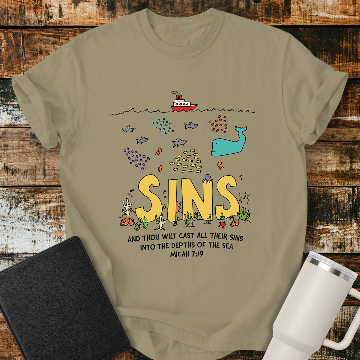 khaki Christian t-shirt featuring a colorful undersea illustration with the word “SINS” resting on the ocean floor, surrounded by fish, a whale, coral, and sea life, with Micah 7:19 below: “And thou wilt cast all their sins into the depths of the sea.”