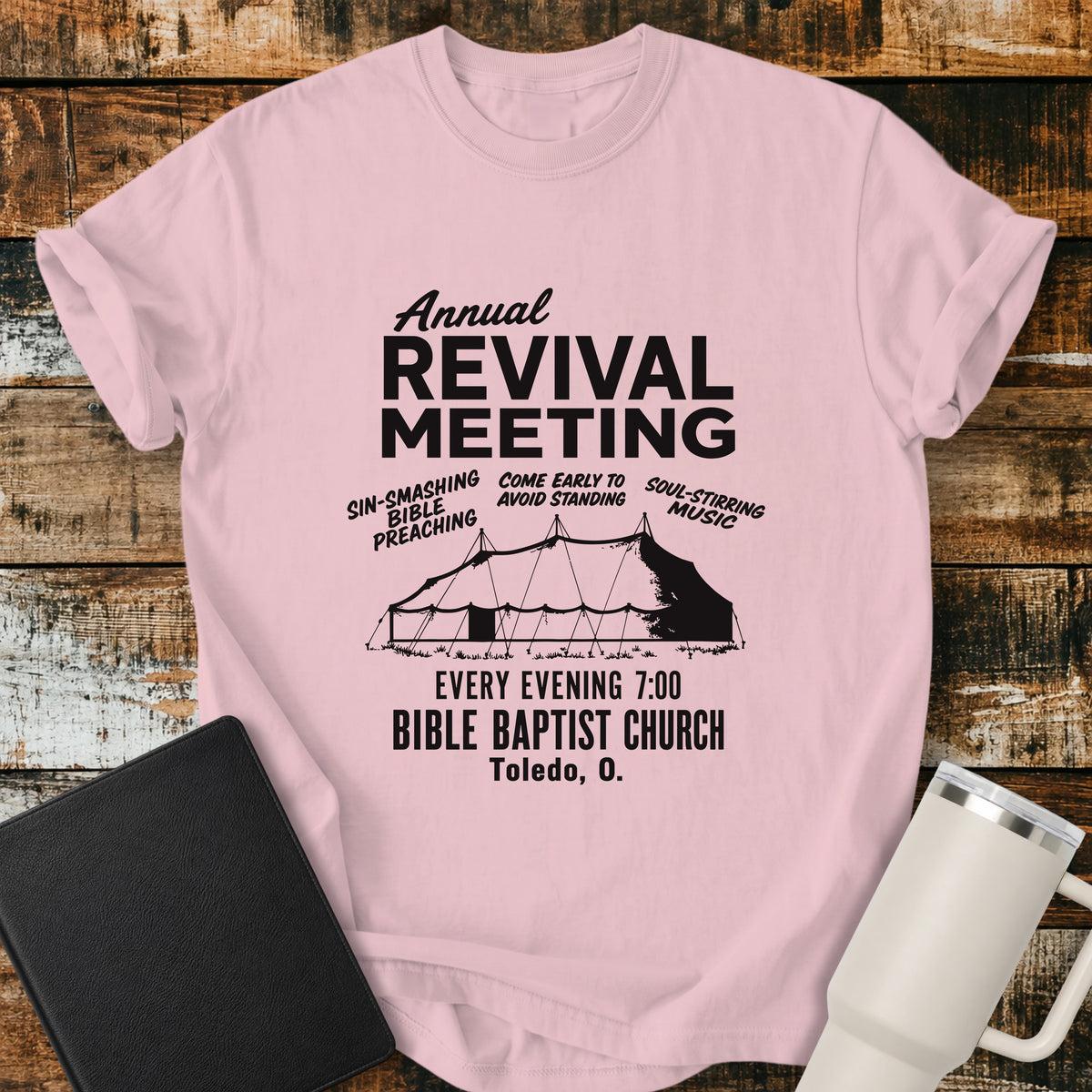 blossom pink flat-lay t-shirt mockup featuring a vintage-inspired church revival design with bold lettering that reads, "Annual Revival Meeting." A classic revival tent is centered below, surrounded by the phrases "Sin-Smashing Bible Preaching," "Come Early to Avoid Standing," and "Soul-Stirring Music." Additional text reads, "Every Evening 7:00," "Bible Baptist Church," and "Toledo, O." The shirt is displayed on a rustic wood plank background alongside a black leather Bible and a white insulated travel mu