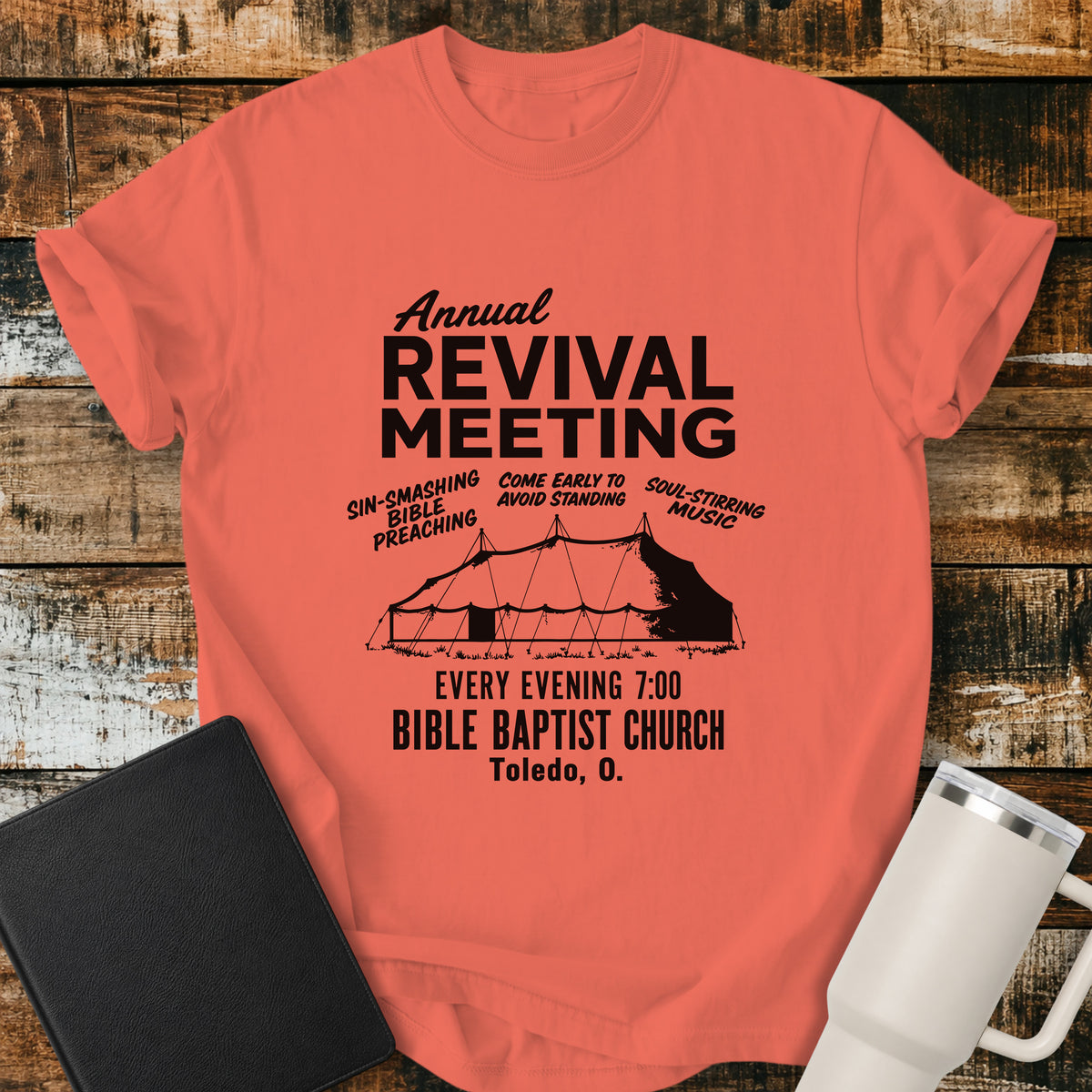 Bright salmon flat-lay t-shirt mockup featuring a vintage-inspired church revival design with bold lettering that reads, "Annual Revival Meeting." A classic revival tent is centered below, surrounded by the phrases "Sin-Smashing Bible Preaching," "Come Early to Avoid Standing," and "Soul-Stirring Music." Additional text reads, "Every Evening 7:00," "Bible Baptist Church," and "Toledo, O." The shirt is displayed on a rustic wood plank background alongside a black leather Bible and a white insulated travel mu