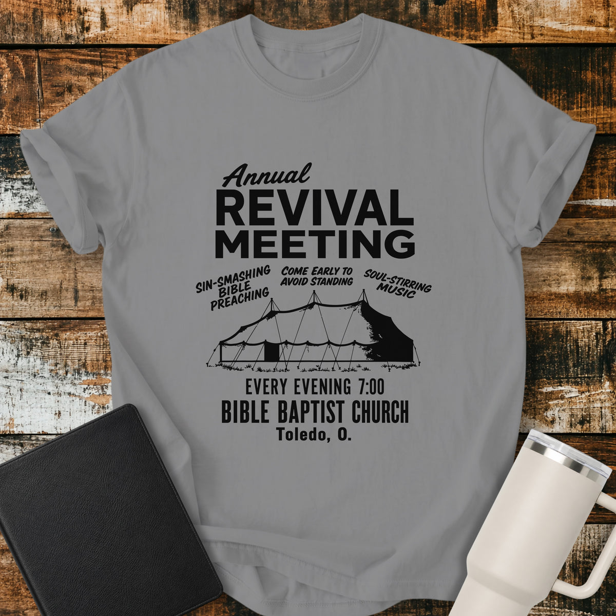 grey flat-lay t-shirt mockup featuring a vintage-inspired church revival design with bold lettering that reads, "Annual Revival Meeting." A classic revival tent is centered below, surrounded by the phrases "Sin-Smashing Bible Preaching," "Come Early to Avoid Standing," and "Soul-Stirring Music." Additional text reads, "Every Evening 7:00," "Bible Baptist Church," and "Toledo, O." The shirt is displayed on a rustic wood plank background alongside a black leather Bible and a white insulated travel mu