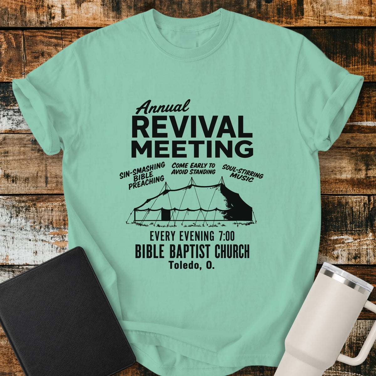 island reef flat-lay t-shirt mockup featuring a vintage-inspired church revival design with bold lettering that reads, "Annual Revival Meeting." A classic revival tent is centered below, surrounded by the phrases "Sin-Smashing Bible Preaching," "Come Early to Avoid Standing," and "Soul-Stirring Music." Additional text reads, "Every Evening 7:00," "Bible Baptist Church," and "Toledo, O." The shirt is displayed on a rustic wood plank background alongside a black leather Bible and a white insulated travel mu