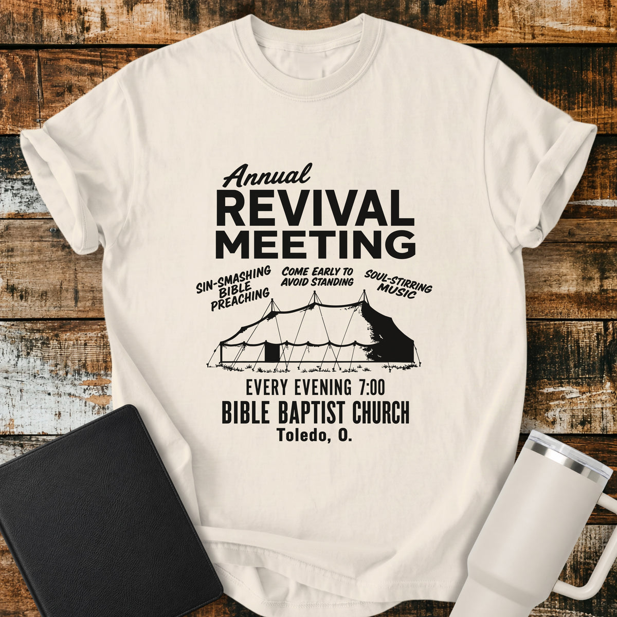 ivory flat-lay t-shirt mockup featuring a vintage-inspired church revival design with bold lettering that reads, "Annual Revival Meeting." A classic revival tent is centered below, surrounded by the phrases "Sin-Smashing Bible Preaching," "Come Early to Avoid Standing," and "Soul-Stirring Music." Additional text reads, "Every Evening 7:00," "Bible Baptist Church," and "Toledo, O." The shirt is displayed on a rustic wood plank background alongside a black leather Bible and a white insulated travel mu