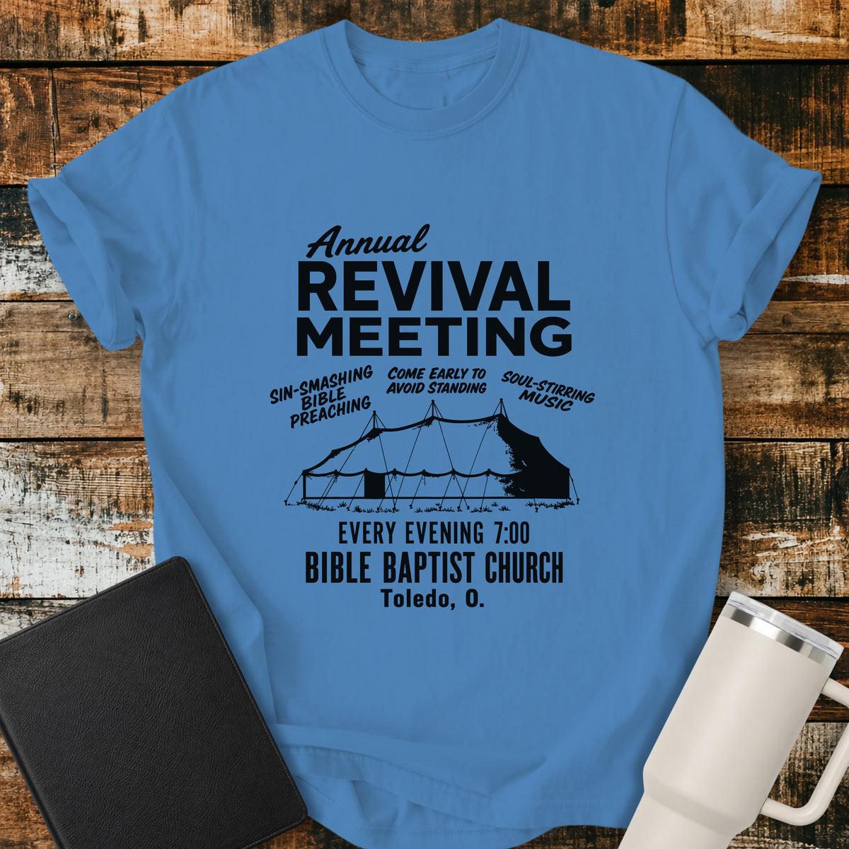 blue caribe flat-lay t-shirt mockup featuring a vintage-inspired church revival design with bold lettering that reads, "Annual Revival Meeting." A classic revival tent is centered below, surrounded by the phrases "Sin-Smashing Bible Preaching," "Come Early to Avoid Standing," and "Soul-Stirring Music." Additional text reads, "Every Evening 7:00," "Bible Baptist Church," and "Toledo, O." The shirt is displayed on a rustic wood plank background alongside a black leather Bible and a white insulated travel mu