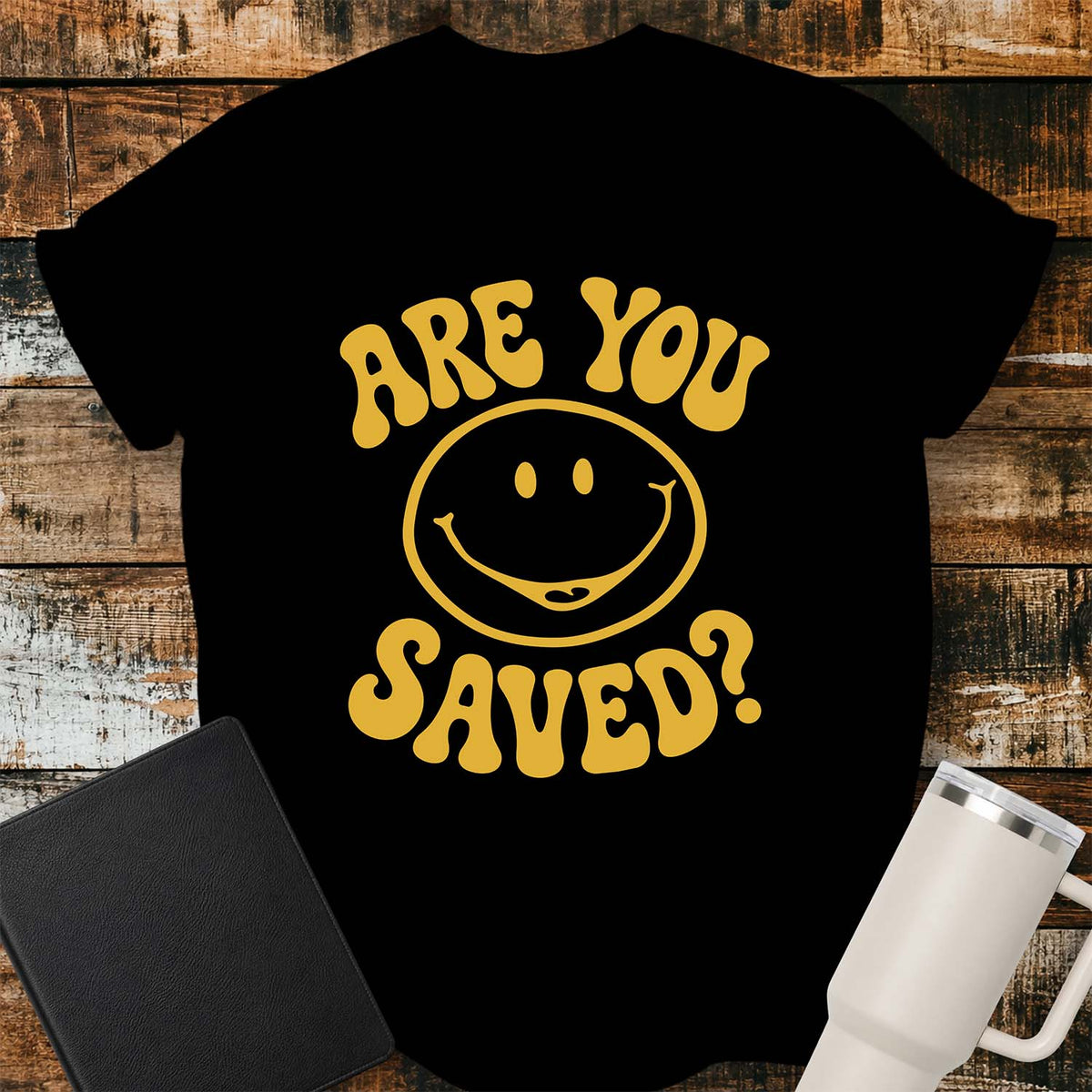 black flat-lay t-shirt mockup featuring a retro groovy yellow smiley face surrounded by bold vintage lettering that reads, "ARE YOU SAVED?" The simple faith-inspired design is centered on the shirt and displayed on a rustic wood plank background with a black leather Bible and a white insulated travel mug.