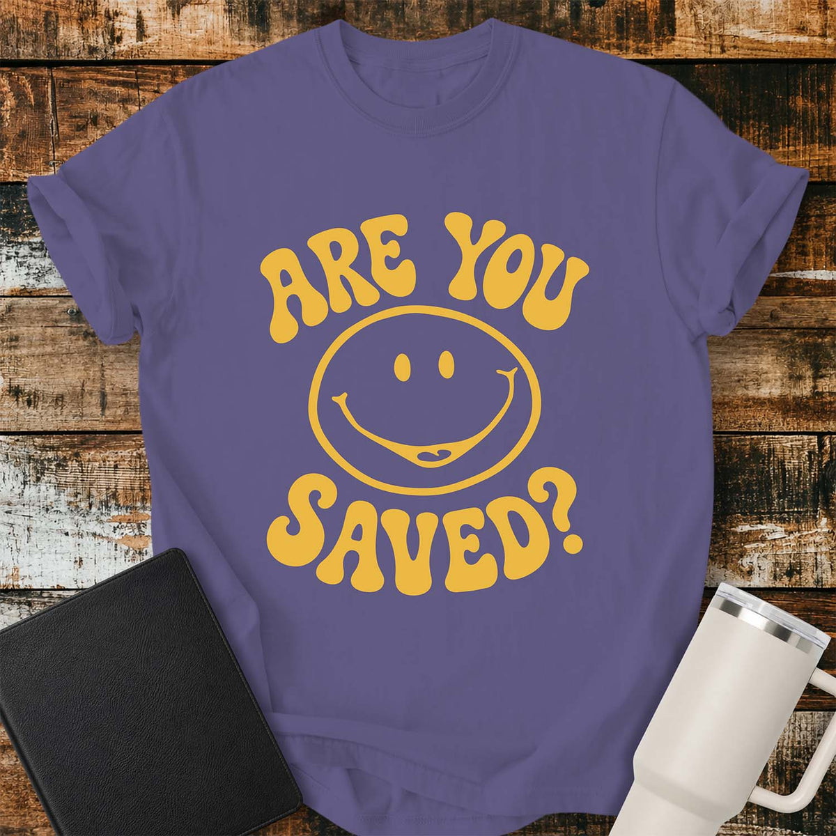 grape flat-lay t-shirt mockup featuring a retro groovy yellow smiley face surrounded by bold vintage lettering that reads, "ARE YOU SAVED?" The simple faith-inspired design is centered on the shirt and displayed on a rustic wood plank background with a black leather Bible and a white insulated travel mug.