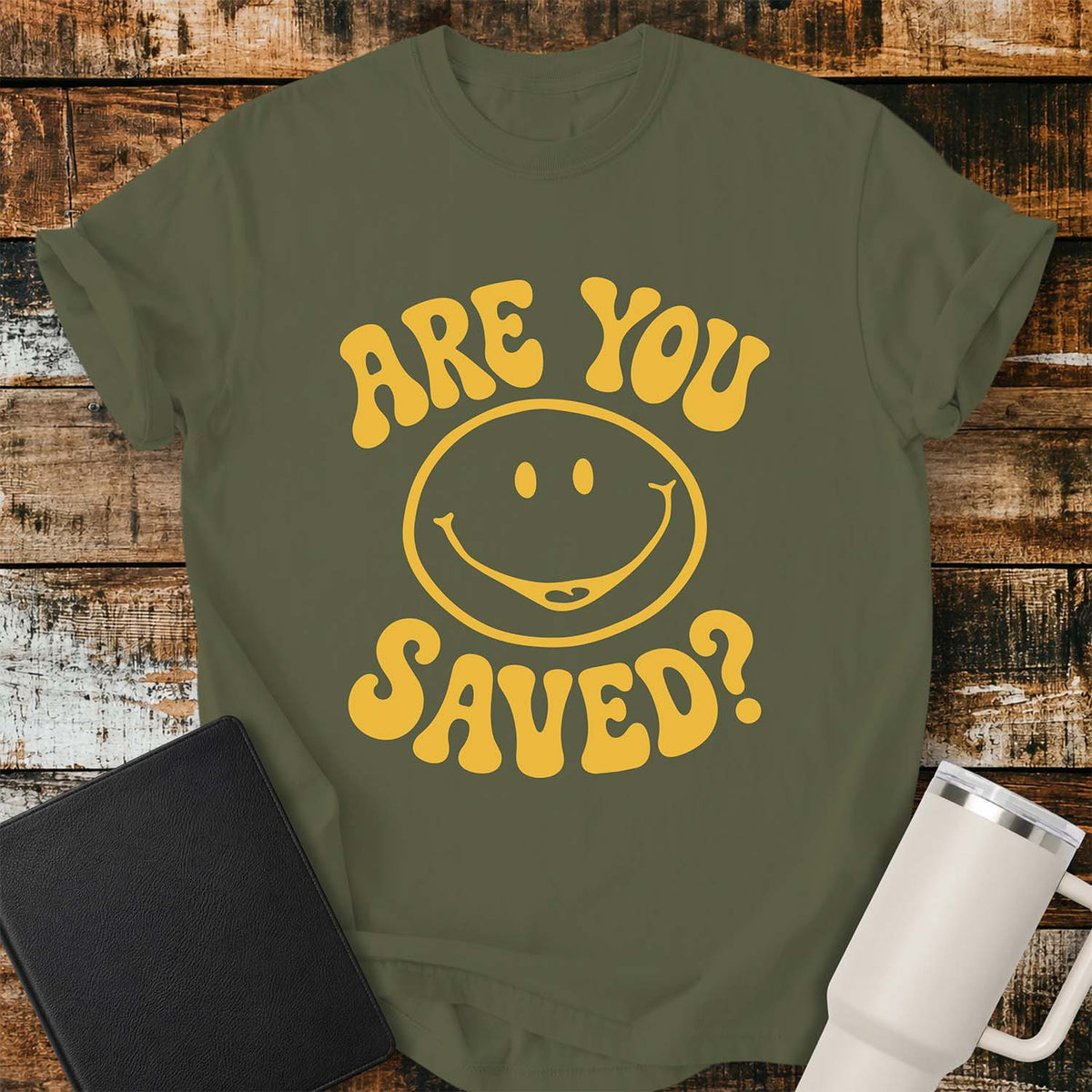 hemp green flat-lay t-shirt mockup featuring a retro groovy yellow smiley face surrounded by bold vintage lettering that reads, "ARE YOU SAVED?" The simple faith-inspired design is centered on the shirt and displayed on a rustic wood plank background with a black leather Bible and a white insulated travel mug.