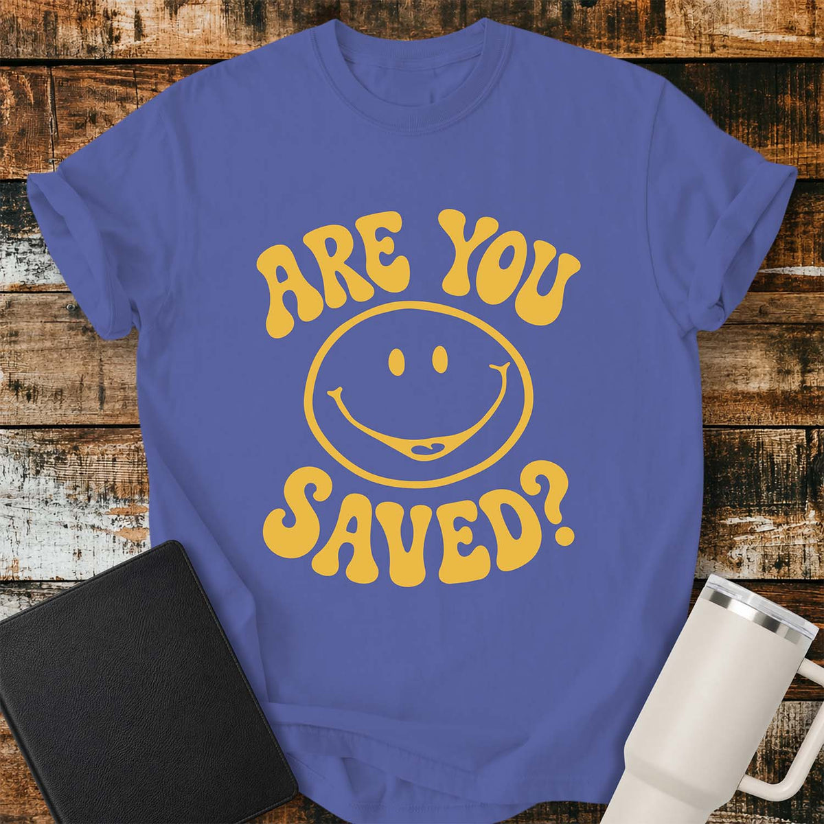 periwinkle flat-lay t-shirt mockup featuring a retro groovy yellow smiley face surrounded by bold vintage lettering that reads, "ARE YOU SAVED?" The simple faith-inspired design is centered on the shirt and displayed on a rustic wood plank background with a black leather Bible and a white insulated travel mug.
