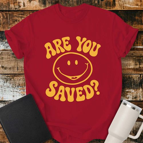 Red flat-lay t-shirt mockup featuring a retro groovy yellow smiley face surrounded by bold vintage lettering that reads, "ARE YOU SAVED?" The simple faith-inspired design is centered on the shirt and displayed on a rustic wood plank background with a black leather Bible and a white insulated travel mug.
