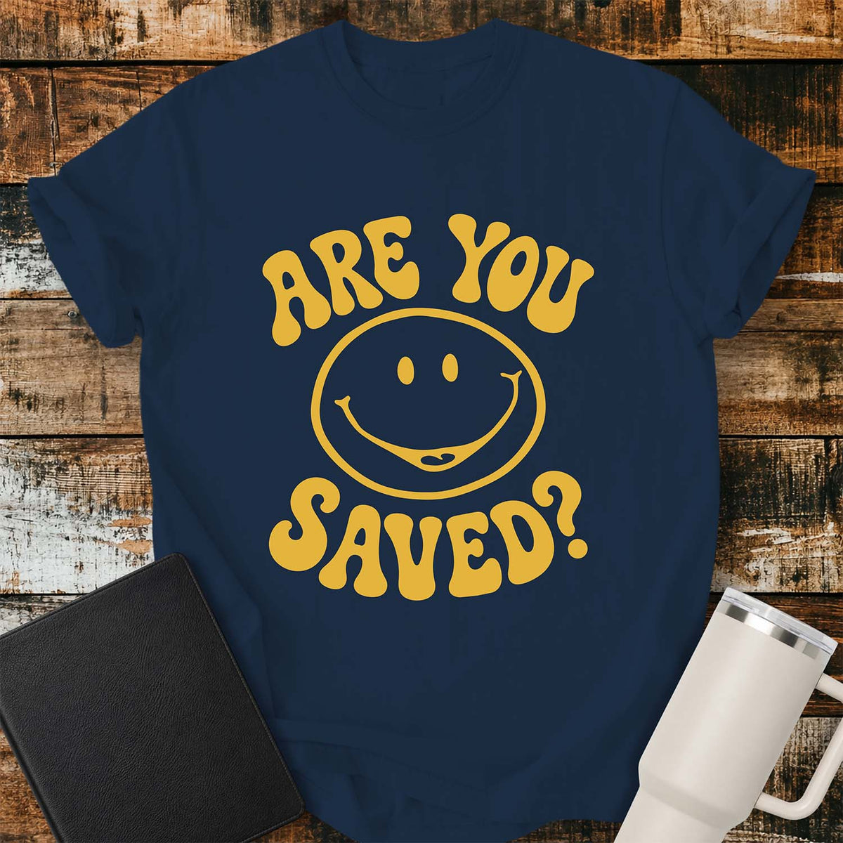 true navy flat-lay t-shirt mockup featuring a retro groovy yellow smiley face surrounded by bold vintage lettering that reads, "ARE YOU SAVED?" The simple faith-inspired design is centered on the shirt and displayed on a rustic wood plank background with a black leather Bible and a white insulated travel mug.