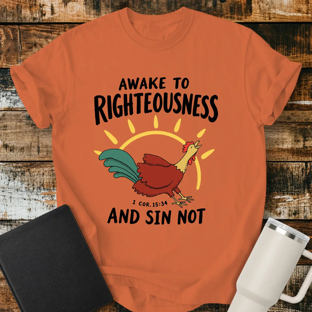 Burnt orange graphic T-shirt mockup featuring a rooster illustration with a rising sun and the phrase “Awake to Righteousness and Sin Not” with a 1 Corinthians 15:34 reference, styled with a black Bible and white tumbler on a rustic wood background.
