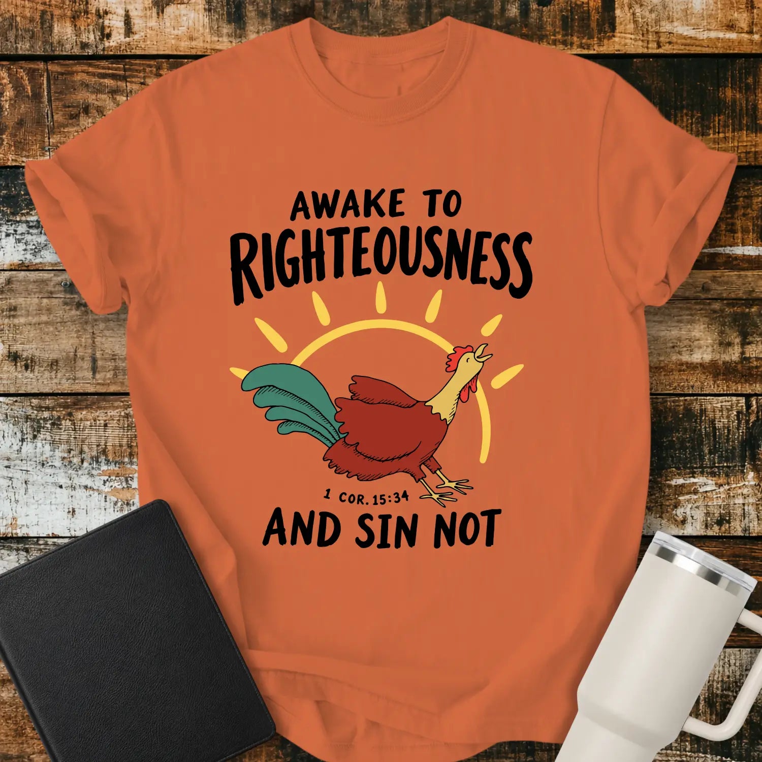 Burnt orange graphic T-shirt mockup featuring a rooster illustration with a rising sun and the phrase “Awake to Righteousness and Sin Not” with a 1 Corinthians 15:34 reference, styled with a black Bible and white tumbler on a rustic wood background.
