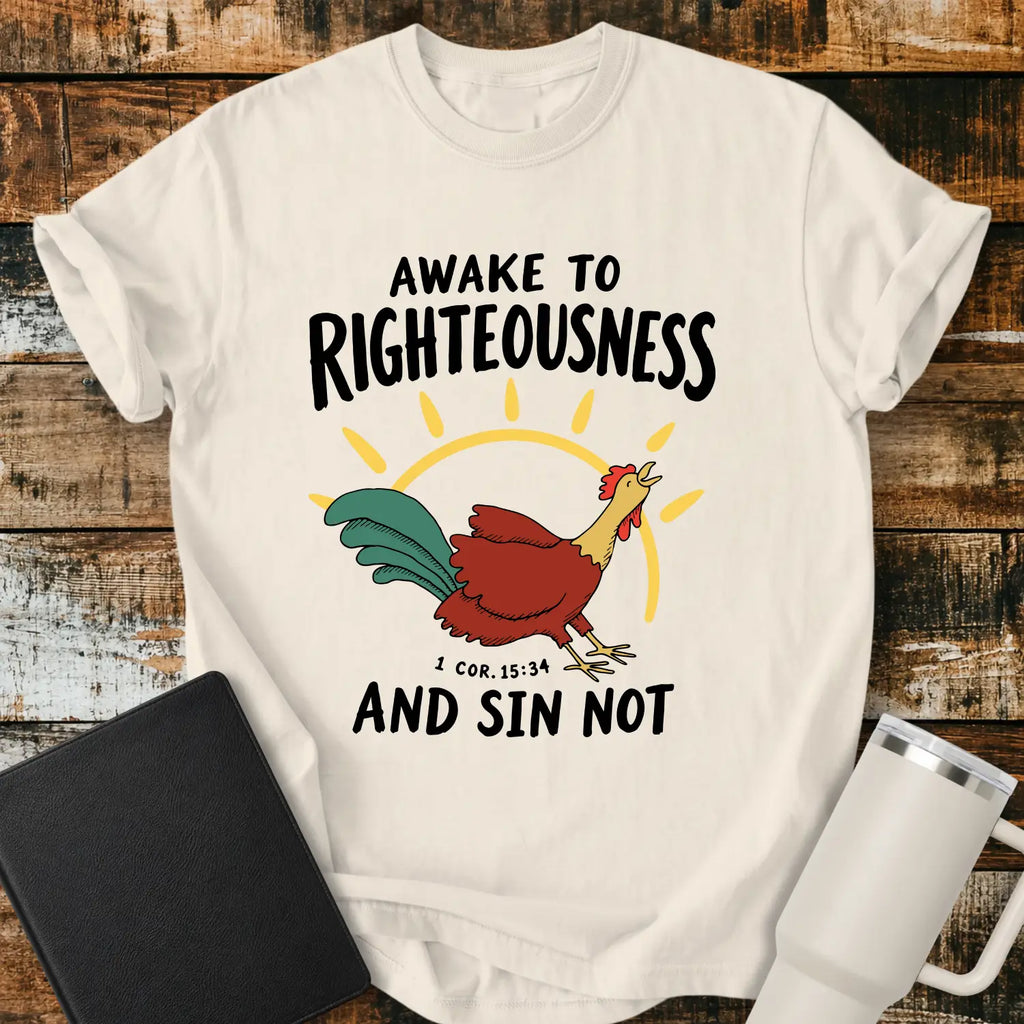 ivory graphic T-shirt mockup featuring a rooster illustration with a rising sun and the phrase “Awake to Righteousness and Sin Not” with a 1 Corinthians 15:34 reference, styled with a black Bible and white tumbler on a rustic wood background.
