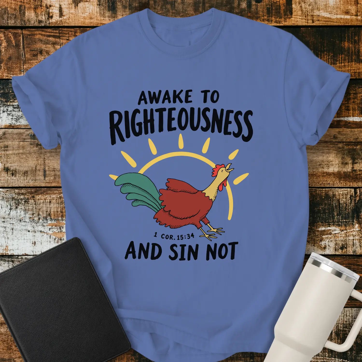 mystic blue graphic T-shirt mockup featuring a rooster illustration with a rising sun and the phrase “Awake to Righteousness and Sin Not” with a 1 Corinthians 15:34 reference, styled with a black Bible and white tumbler on a rustic wood background.
