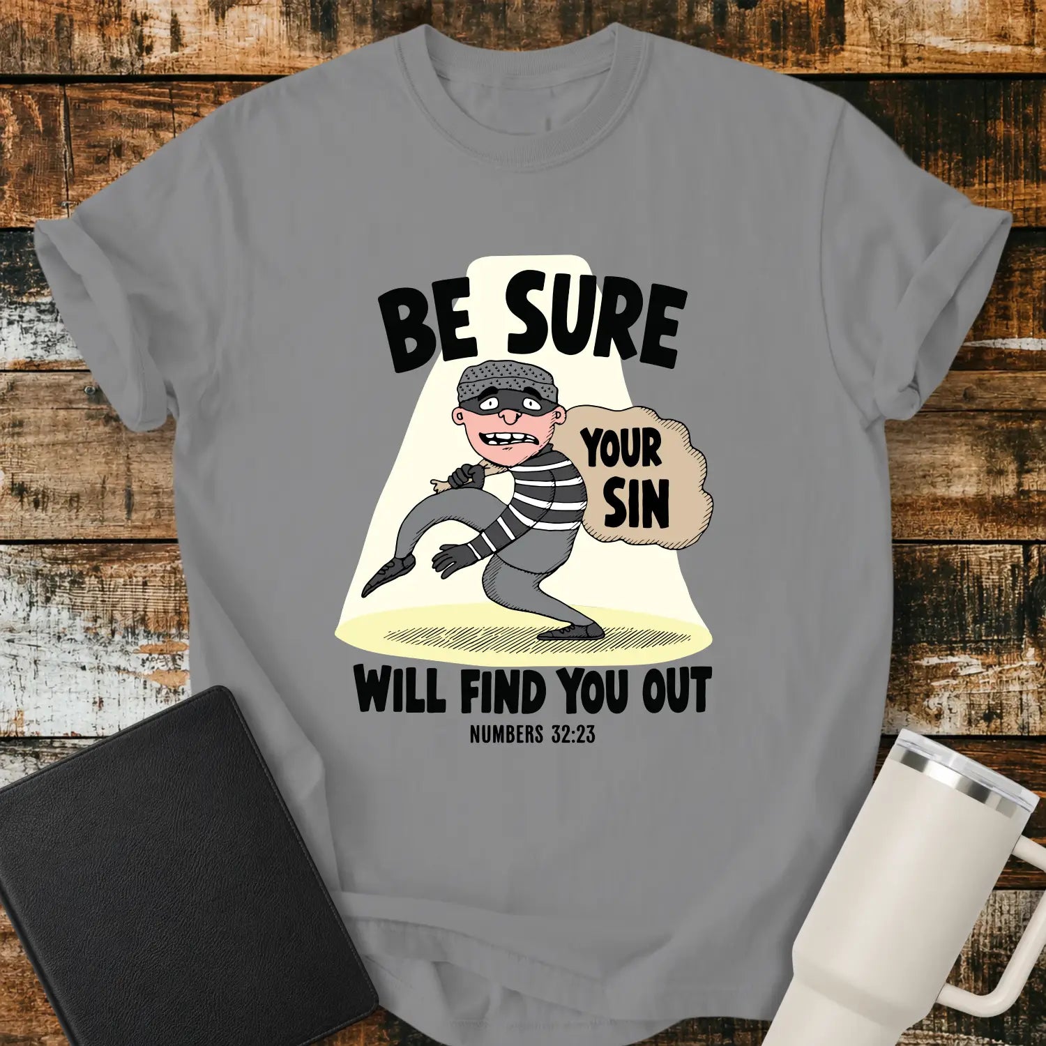 Grey T-shirt featuring a humorous Christian illustration of a startled cartoon burglar caught in a spotlight while carrying a sack labeled “YOUR SIN.” Bold black text reads, “BE SURE YOUR SIN WILL FIND YOU OUT,” with the Bible reference “NUMBERS 32:23” below. The shirt is displayed on a rustic wood plank background alongside a black Bible and a white insulated tumbler.