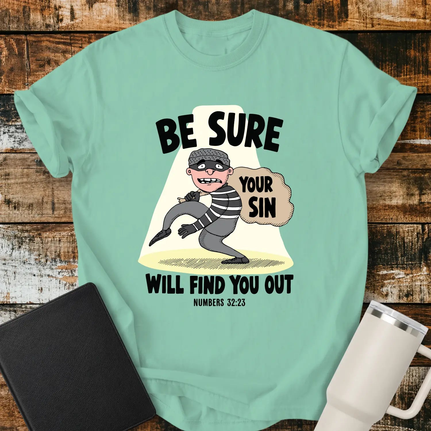 Island reef green T-shirt featuring a humorous Christian illustration of a startled cartoon burglar caught in a spotlight while carrying a sack labeled “YOUR SIN.” Bold black text reads, “BE SURE YOUR SIN WILL FIND YOU OUT,” with the Bible reference “NUMBERS 32:23” below. The shirt is displayed on a rustic wood plank background alongside a black Bible and a white insulated tumbler.