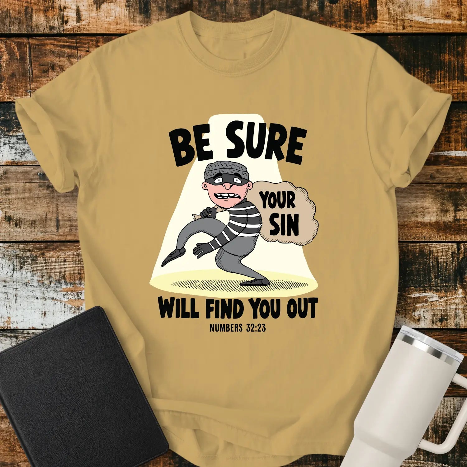 Mustard yellow T-shirt featuring a humorous Christian illustration of a startled cartoon burglar caught in a spotlight while carrying a sack labeled “YOUR SIN.” Bold black text reads, “BE SURE YOUR SIN WILL FIND YOU OUT,” with the Bible reference “NUMBERS 32:23” below. The shirt is displayed on a rustic wood plank background alongside a black Bible and a white insulated tumbler.