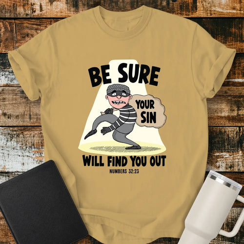 Mustard yellow T-shirt featuring a humorous Christian illustration of a startled cartoon burglar caught in a spotlight while carrying a sack labeled “YOUR SIN.” Bold black text reads, “BE SURE YOUR SIN WILL FIND YOU OUT,” with the Bible reference “NUMBERS 32:23” below. The shirt is displayed on a rustic wood plank background alongside a black Bible and a white insulated tumbler.