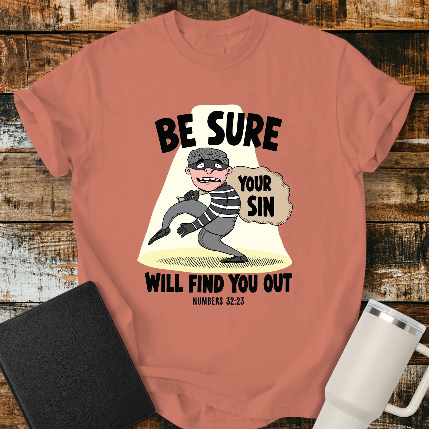 Terracotta T-shirt featuring a humorous Christian illustration of a startled cartoon burglar caught in a spotlight while carrying a sack labeled “YOUR SIN.” Bold black text reads, “BE SURE YOUR SIN WILL FIND YOU OUT,” with the Bible reference “NUMBERS 32:23” below. The shirt is displayed on a rustic wood plank background alongside a black Bible and a white insulated tumbler.