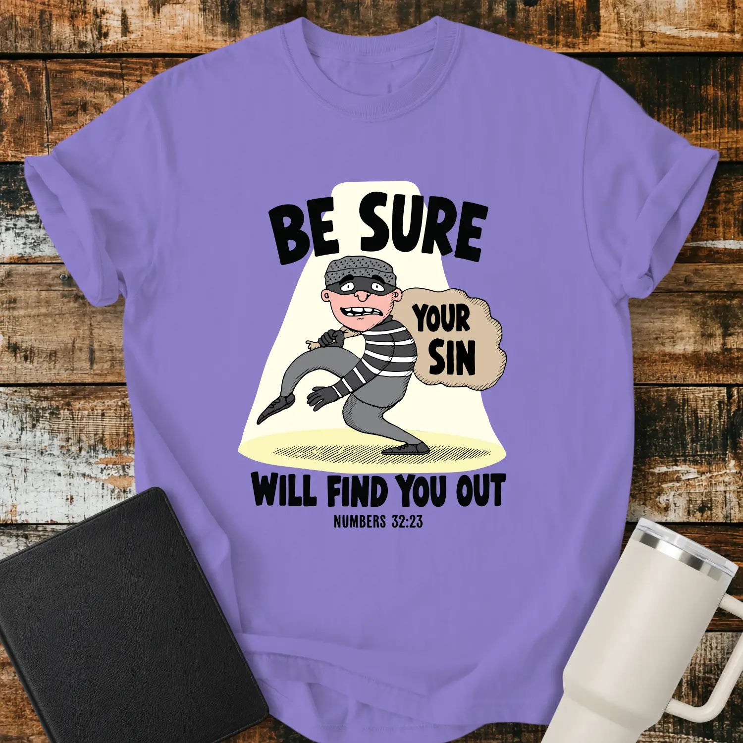 Violet T-shirt featuring a humorous Christian illustration of a startled cartoon burglar caught in a spotlight while carrying a sack labeled “YOUR SIN.” Bold black text reads, “BE SURE YOUR SIN WILL FIND YOU OUT,” with the Bible reference “NUMBERS 32:23” below. The shirt is displayed on a rustic wood plank background alongside a black Bible and a white insulated tumbler.