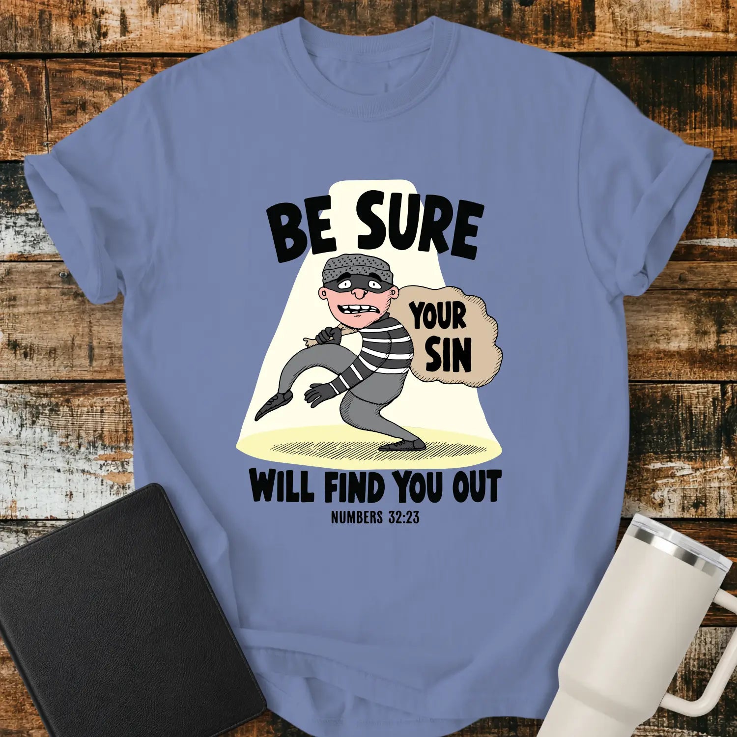 Washed denim blue T-shirt featuring a humorous Christian illustration of a startled cartoon burglar caught in a spotlight while carrying a sack labeled “YOUR SIN.” Bold black text reads, “BE SURE YOUR SIN WILL FIND YOU OUT,” with the Bible reference “NUMBERS 32:23” below. The shirt is displayed on a rustic wood plank background alongside a black Bible and a white insulated tumbler.