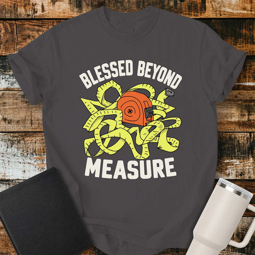 graphite T-shirt featuring a cheerful illustration of a tangled yellow measuring tape wrapped around an orange tape measure. Large white lettering reads “BLESSED BEYOND” above the graphic and “MEASURE” below. The design uses a bold, retro-inspired color palette. The shirt is displayed on a rustic wood plank background with a black Bible on the left and a white insulated tumbler on the right.