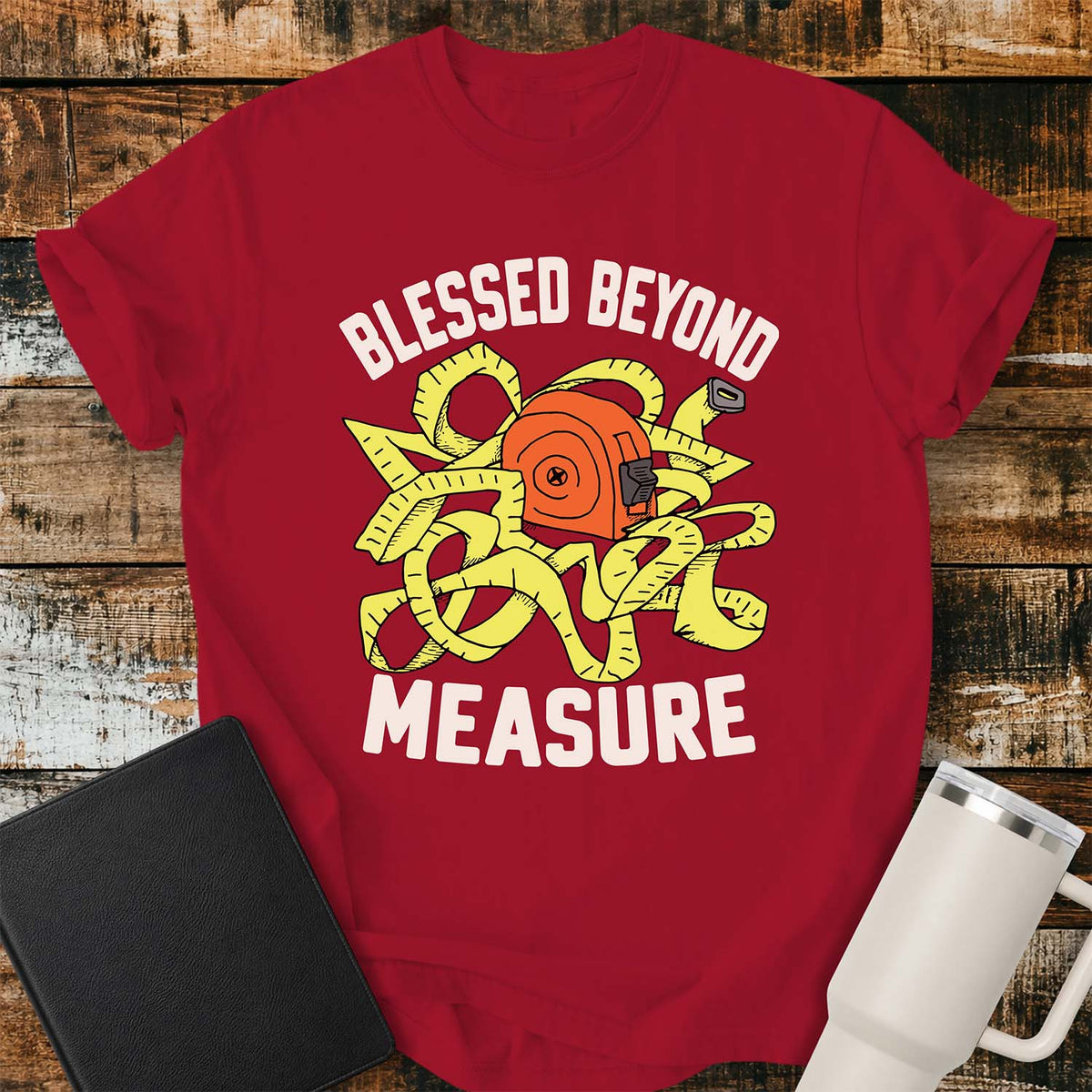 Red T-shirt featuring a cheerful illustration of a tangled yellow measuring tape wrapped around an orange tape measure. Large white lettering reads “BLESSED BEYOND” above the graphic and “MEASURE” below. The design uses a bold, retro-inspired color palette. The shirt is displayed on a rustic wood plank background with a black Bible on the left and a white insulated tumbler on the right.