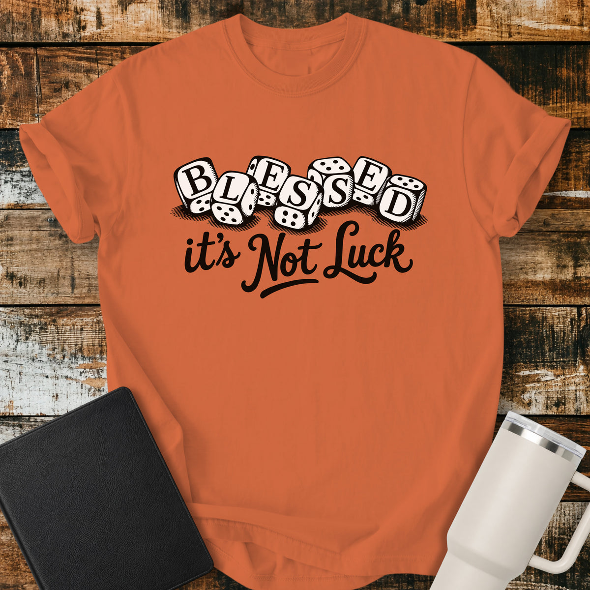 burnt orange flat-lay t-shirt mockup featuring seven white dice spelling the word "BLESSED" above the phrase, "it's Not Luck," in bold black script. The faith-inspired design emphasizes that blessings come from God rather than chance. The shirt is displayed on a rustic wood plank background alongside a black leather Bible and a white insulated travel mug.