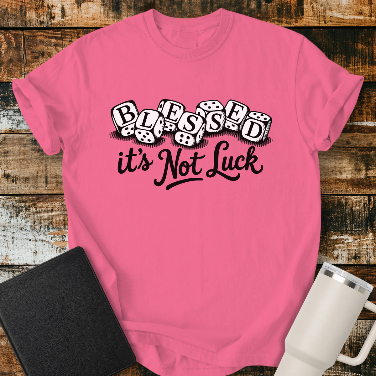 crunchberry pink flat-lay t-shirt mockup featuring seven white dice spelling the word "BLESSED" above the phrase, "it's Not Luck," in bold black script. The faith-inspired design emphasizes that blessings come from God rather than chance. The shirt is displayed on a rustic wood plank background alongside a black leather Bible and a white insulated travel mug.