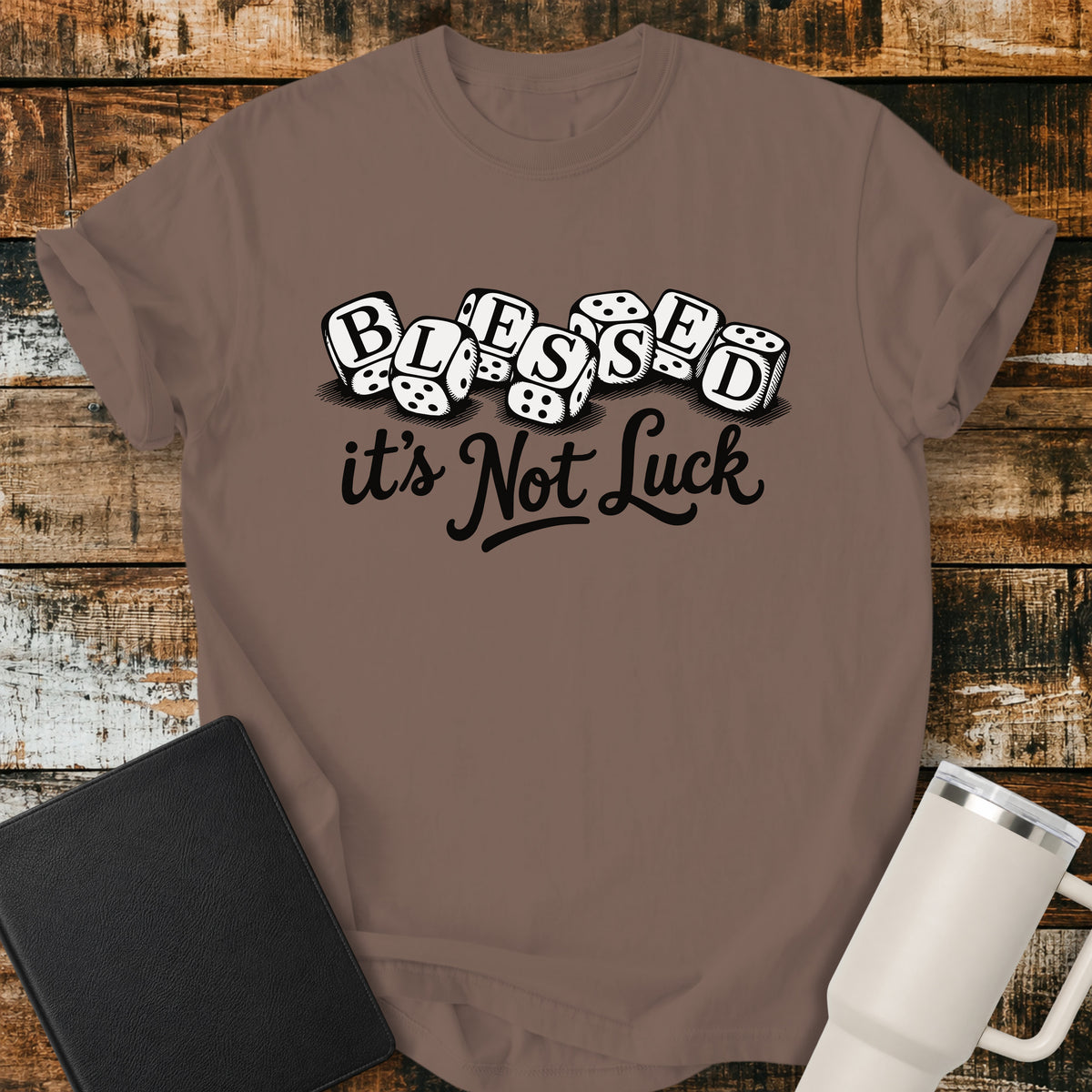 espresso brown flat-lay t-shirt mockup featuring seven white dice spelling the word "BLESSED" above the phrase, "it's Not Luck," in bold black script. The faith-inspired design emphasizes that blessings come from God rather than chance. The shirt is displayed on a rustic wood plank background alongside a black leather Bible and a white insulated travel mug.