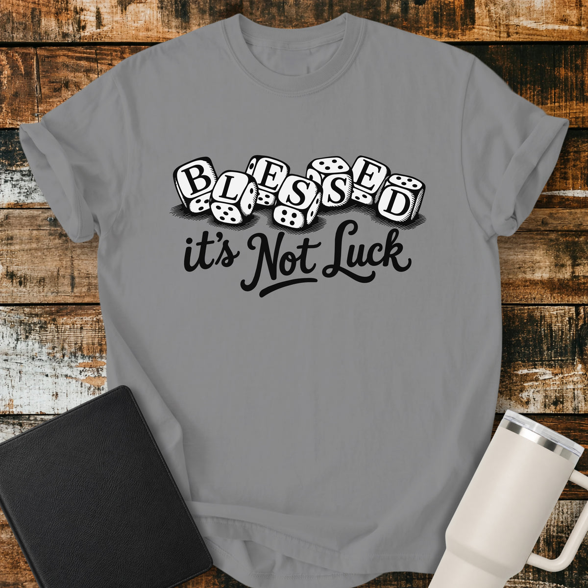 grey flat-lay t-shirt mockup featuring seven white dice spelling the word "BLESSED" above the phrase, "it's Not Luck," in bold black script. The faith-inspired design emphasizes that blessings come from God rather than chance. The shirt is displayed on a rustic wood plank background alongside a black leather Bible and a white insulated travel mug.