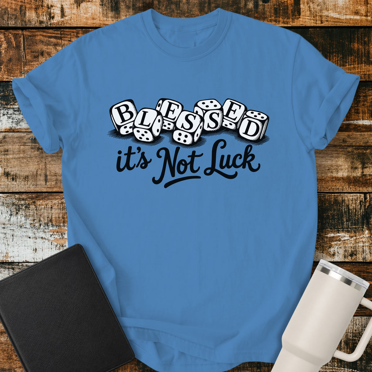 Royal caribe blue flat-lay t-shirt mockup featuring seven white dice spelling the word "BLESSED" above the phrase, "it's Not Luck," in bold black script. The faith-inspired design emphasizes that blessings come from God rather than chance. The shirt is displayed on a rustic wood plank background alongside a black leather Bible and a white insulated travel mug.