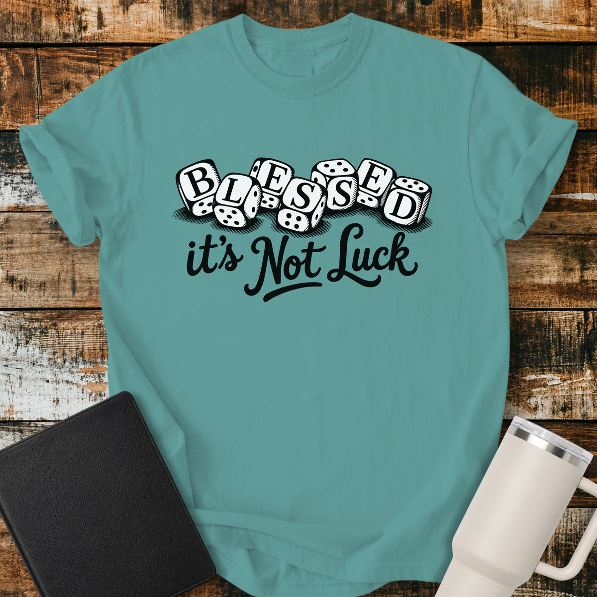 seafoam green flat-lay t-shirt mockup featuring seven white dice spelling the word "BLESSED" above the phrase, "it's Not Luck," in bold black script. The faith-inspired design emphasizes that blessings come from God rather than chance. The shirt is displayed on a rustic wood plank background alongside a black leather Bible and a white insulated travel mug.