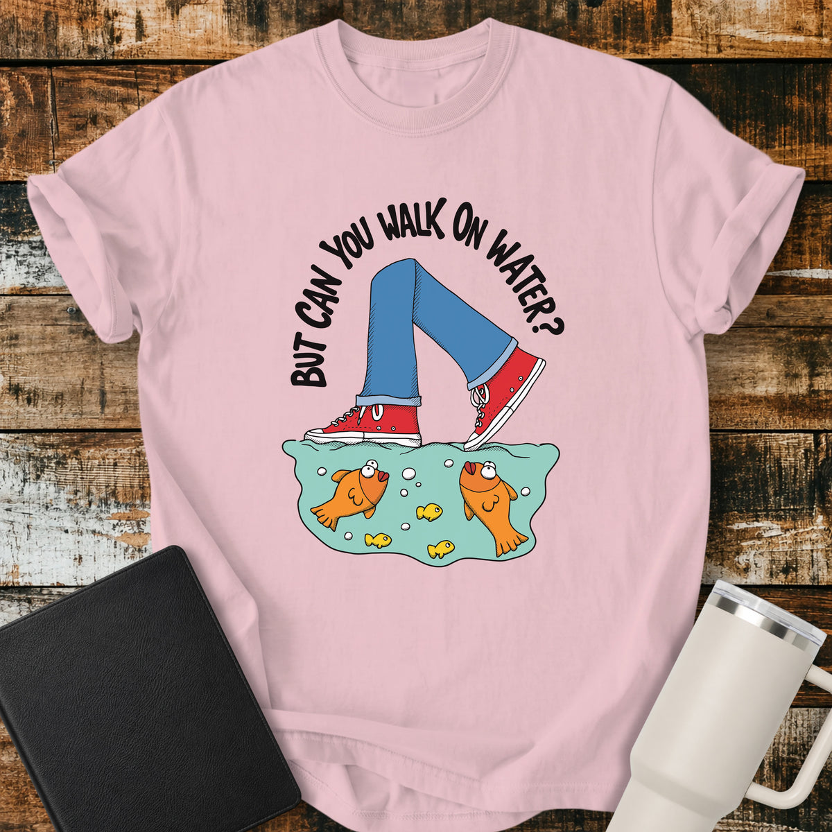 blossom pink flat-lay t-shirt mockup featuring a playful illustration of a person wearing red sneakers walking across the surface of the water above cartoon goldfish. Curved white lettering reads, "BUT CAN YOU WALK ON WATER?" The faith-inspired design references Jesus walking on water and is displayed on a rustic wood plank background alongside a black leather Bible and a white insulated travel mug.