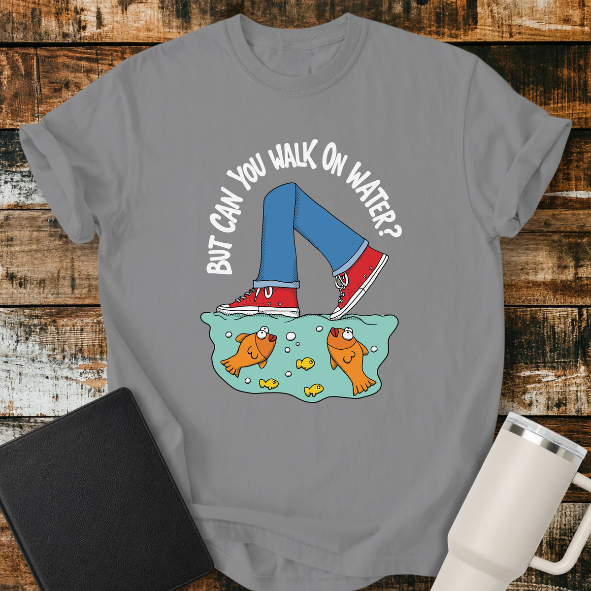 grey flat-lay t-shirt mockup featuring a playful illustration of a person wearing red sneakers walking across the surface of the water above cartoon goldfish. Curved white lettering reads, "BUT CAN YOU WALK ON WATER?" The faith-inspired design references Jesus walking on water and is displayed on a rustic wood plank background alongside a black leather Bible and a white insulated travel mug.
