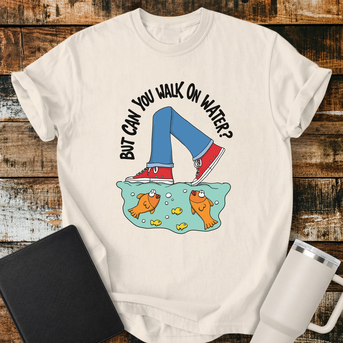 ivory flat-lay t-shirt mockup featuring a playful illustration of a person wearing red sneakers walking across the surface of the water above cartoon goldfish. Curved white lettering reads, "BUT CAN YOU WALK ON WATER?" The faith-inspired design references Jesus walking on water and is displayed on a rustic wood plank background alongside a black leather Bible and a white insulated travel mug.