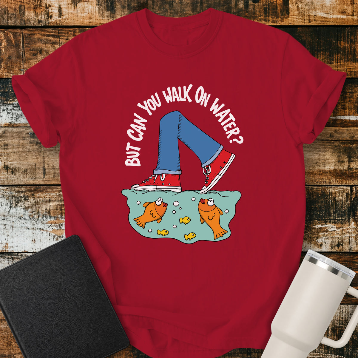 red flat-lay t-shirt mockup featuring a playful illustration of a person wearing red sneakers walking across the surface of the water above cartoon goldfish. Curved white lettering reads, "BUT CAN YOU WALK ON WATER?" The faith-inspired design references Jesus walking on water and is displayed on a rustic wood plank background alongside a black leather Bible and a white insulated travel mug.