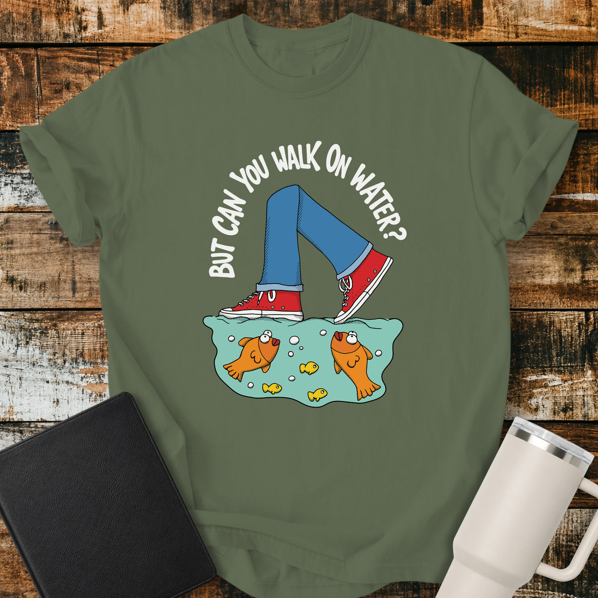 Sage green flat-lay t-shirt mockup featuring a playful illustration of a person wearing red sneakers walking across the surface of the water above cartoon goldfish. Curved white lettering reads, "BUT CAN YOU WALK ON WATER?" The faith-inspired design references Jesus walking on water and is displayed on a rustic wood plank background alongside a black leather Bible and a white insulated travel mug.