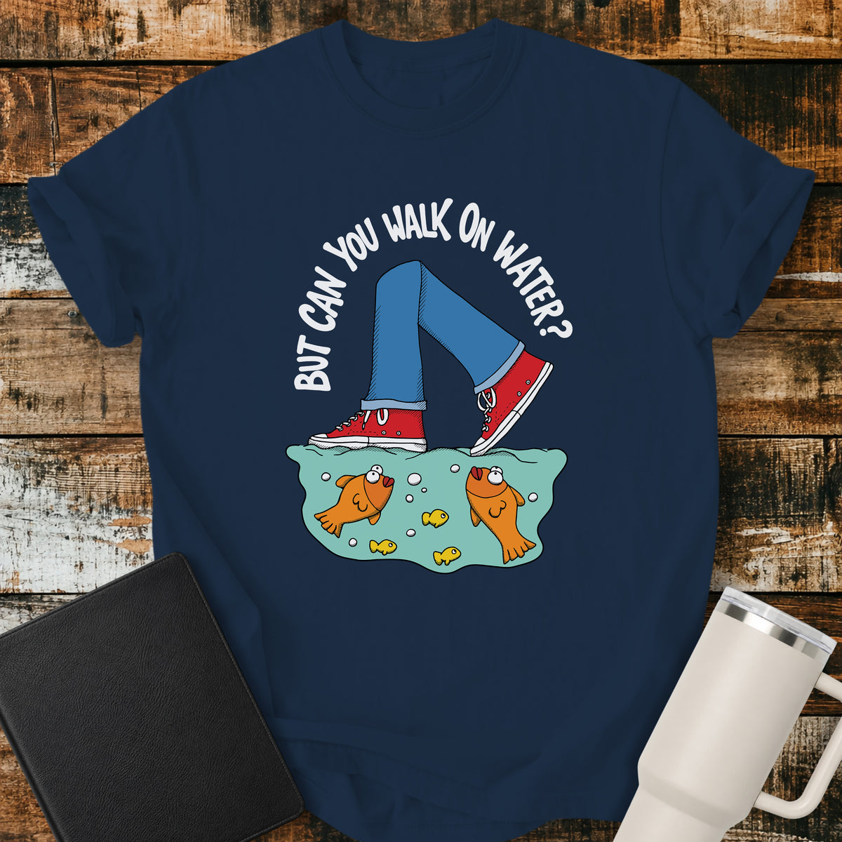 true navy blue flat-lay t-shirt mockup featuring a playful illustration of a person wearing red sneakers walking across the surface of the water above cartoon goldfish. Curved white lettering reads, "BUT CAN YOU WALK ON WATER?" The faith-inspired design references Jesus walking on water and is displayed on a rustic wood plank background alongside a black leather Bible and a white insulated travel mug.