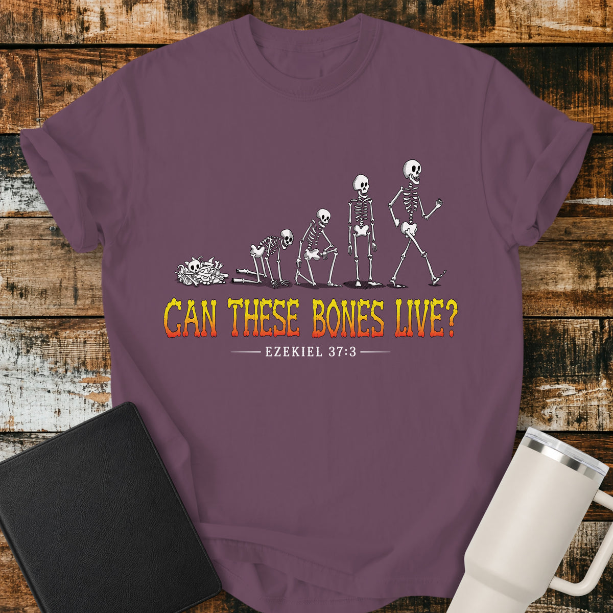 berry flat-lay t-shirt mockup featuring a striking illustration of dry bones transforming into a walking skeleton, inspired by Ezekiel's vision of the valley of dry bones. Bold distressed lettering in a yellow-to-red gradient reads, "CAN THESE BONES LIVE?" with the Bible reference "EZEKIEL 37:3" below. The shirt is displayed on a rustic wood plank background alongside a black leather Bible and a white insulated travel mug.