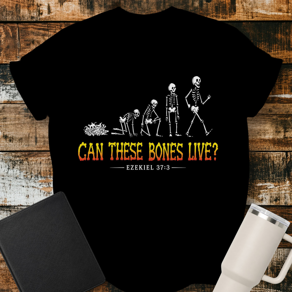 Black flat-lay t-shirt mockup featuring a striking illustration of dry bones transforming into a walking skeleton, inspired by Ezekiel's vision of the valley of dry bones. Bold distressed lettering in a yellow-to-red gradient reads, "CAN THESE BONES LIVE?" with the Bible reference "EZEKIEL 37:3" below. The shirt is displayed on a rustic wood plank background alongside a black leather Bible and a white insulated travel mug.