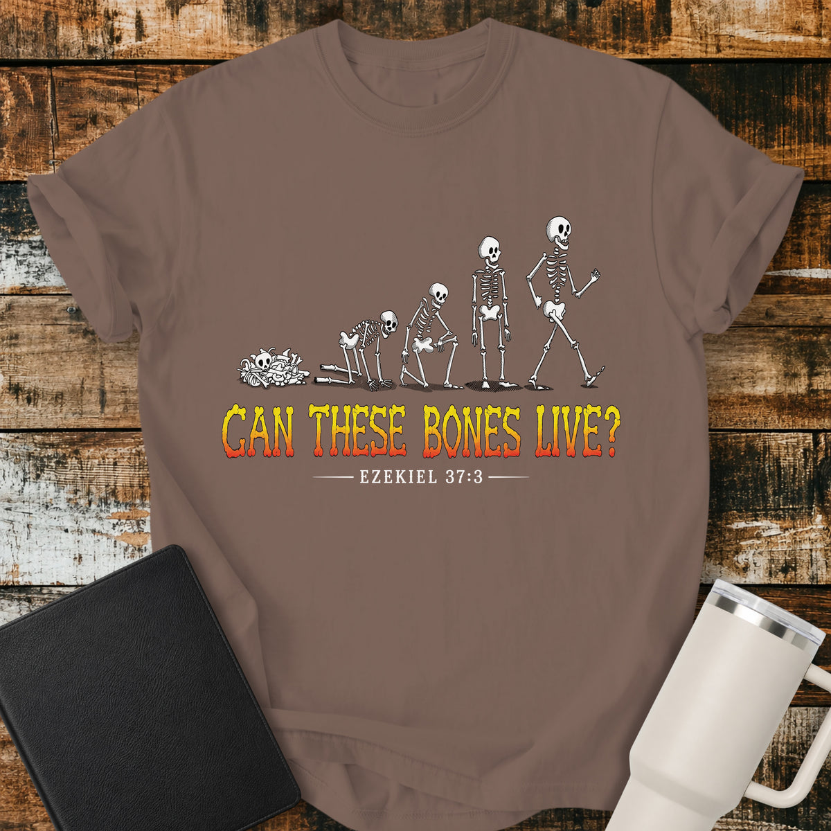espresso brown flat-lay t-shirt mockup featuring a striking illustration of dry bones transforming into a walking skeleton, inspired by Ezekiel's vision of the valley of dry bones. Bold distressed lettering in a yellow-to-red gradient reads, "CAN THESE BONES LIVE?" with the Bible reference "EZEKIEL 37:3" below. The shirt is displayed on a rustic wood plank background alongside a black leather Bible and a white insulated travel mug.